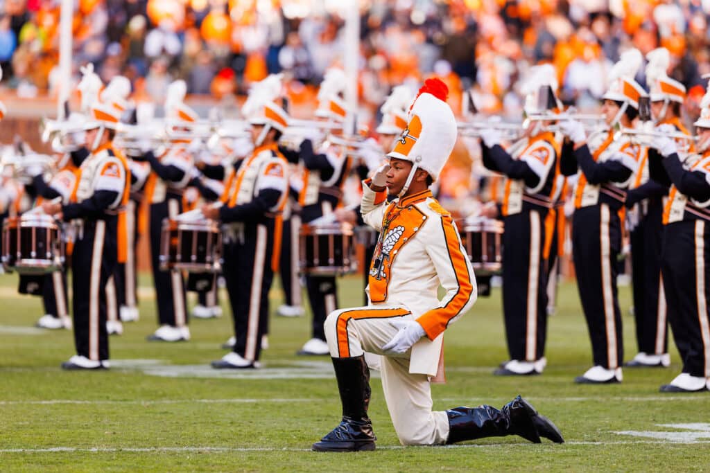 Drum major salutes on a football field with marching band in the background.
