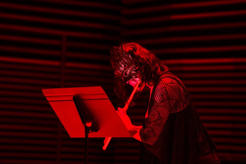 Woman playing a flute on stage with a mask on in red lighting.