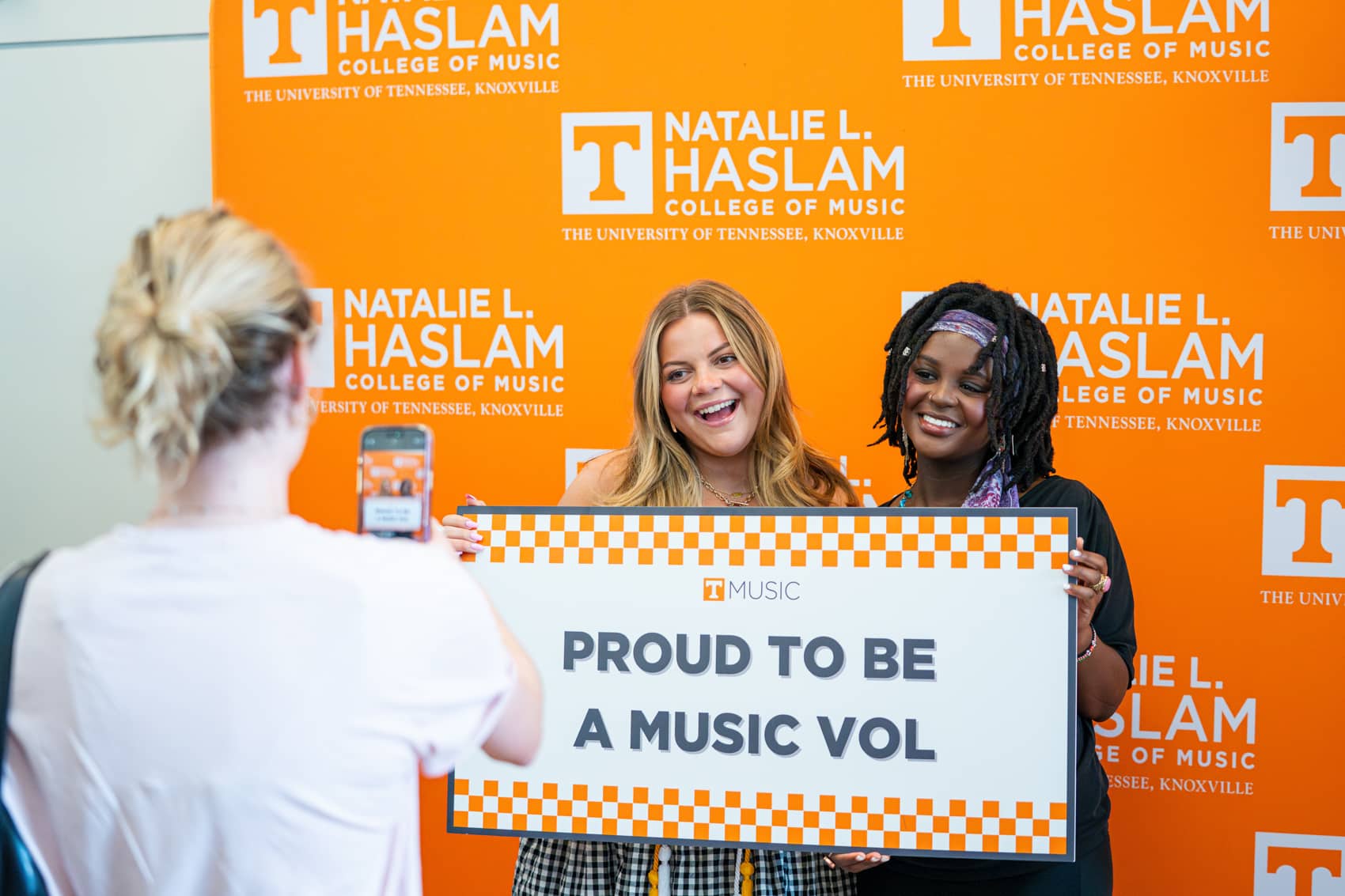 Two women holding "Proud to be a Music Vol" sign in front of orange backdrop