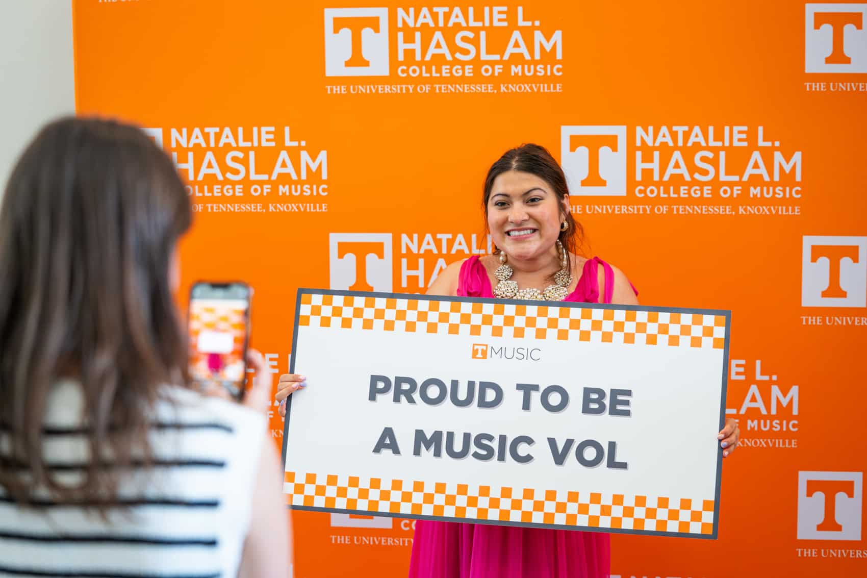 Female student in front of orange back drop holding  a sign that says "Proud to be a Music Vol"