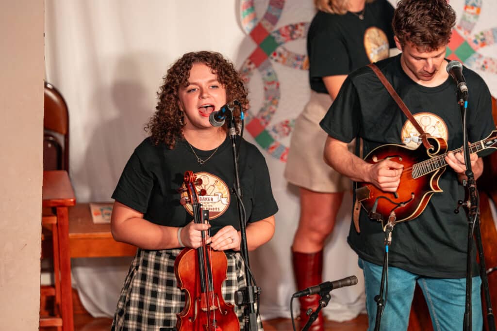 Female student singing on stage holding a fiddle while male student plays guitar next to her.