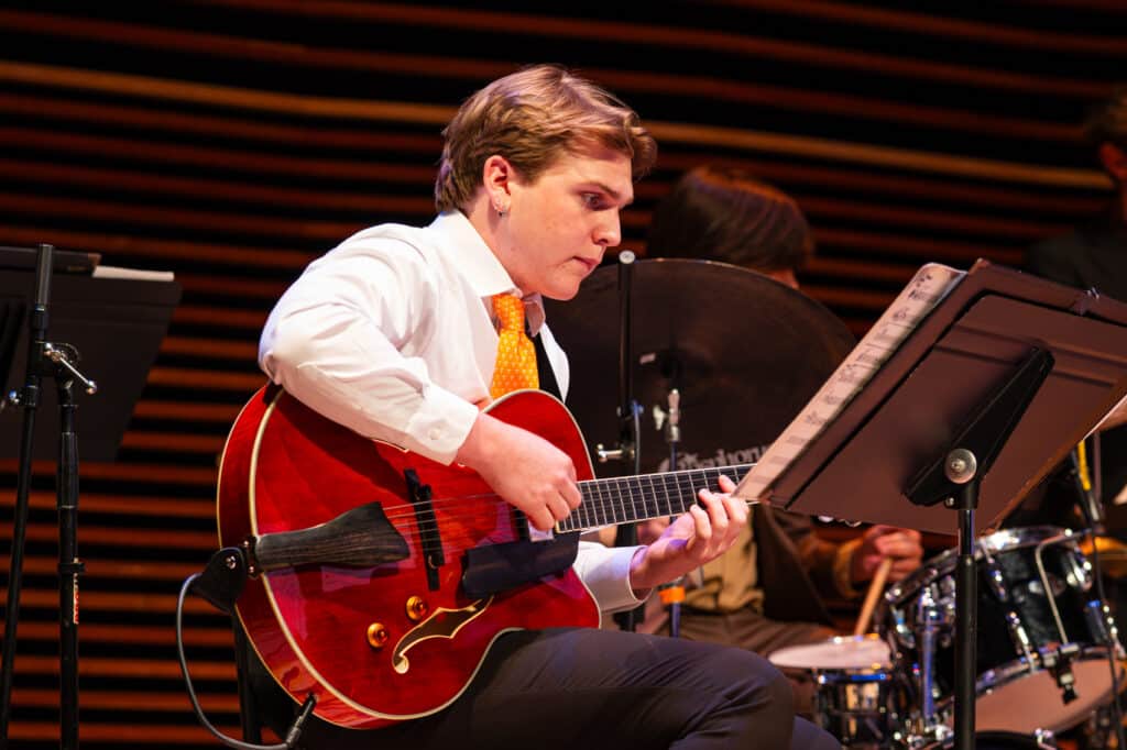 Man on stage looking at sheet music while he plays the guitar.