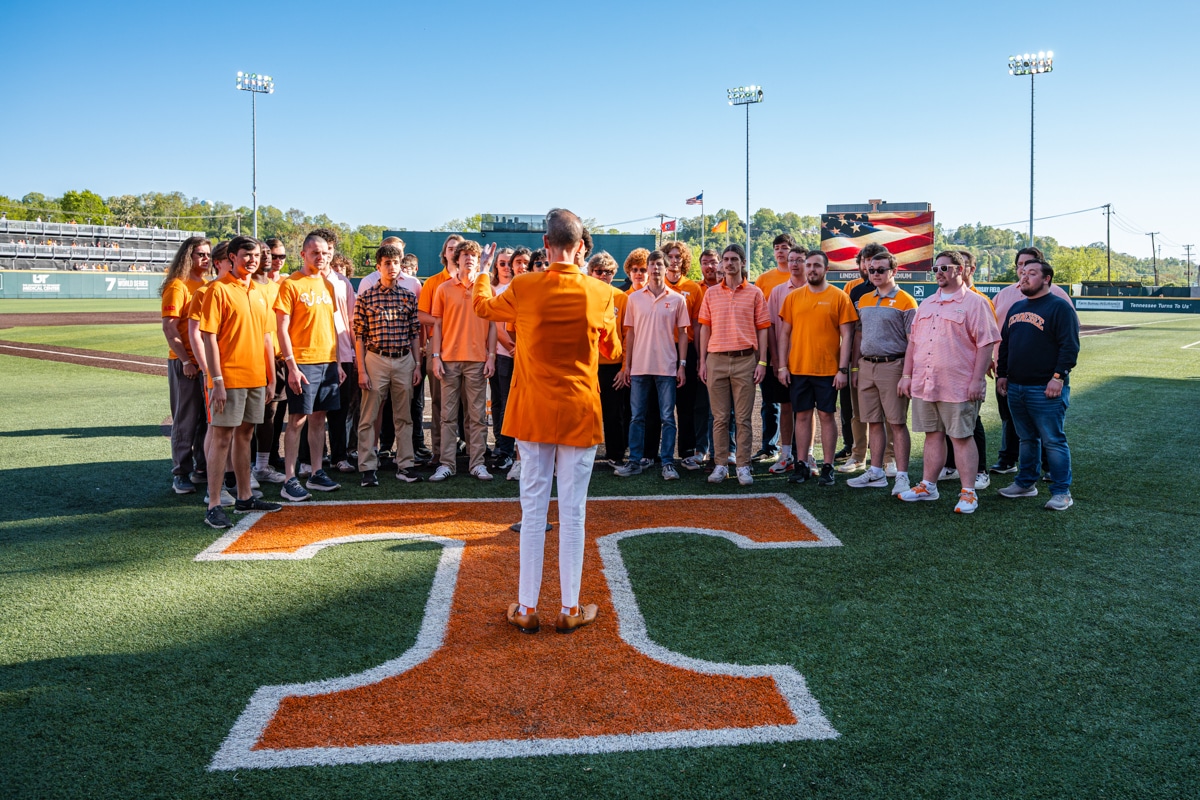 Male conductor in orange suit leads male chorale group on baseball field