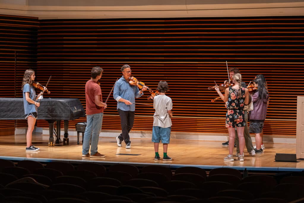 Group of string students listening to a male instructor on stage.