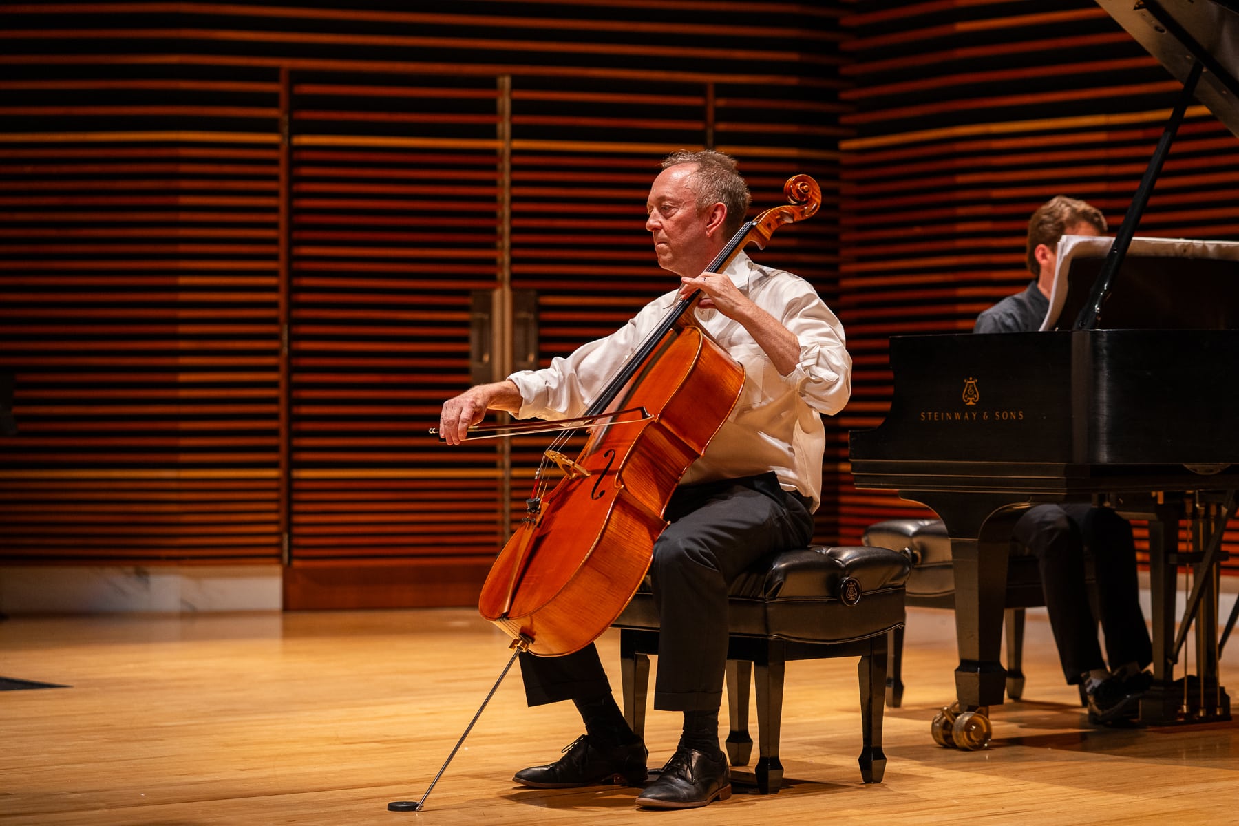 Man playing cello on stage with pianist in the background.