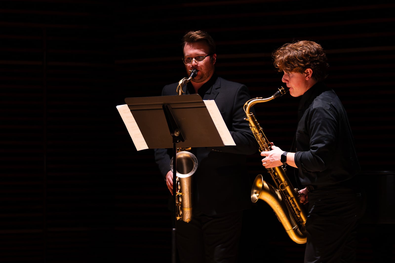 Two saxophone players on a dark stage.