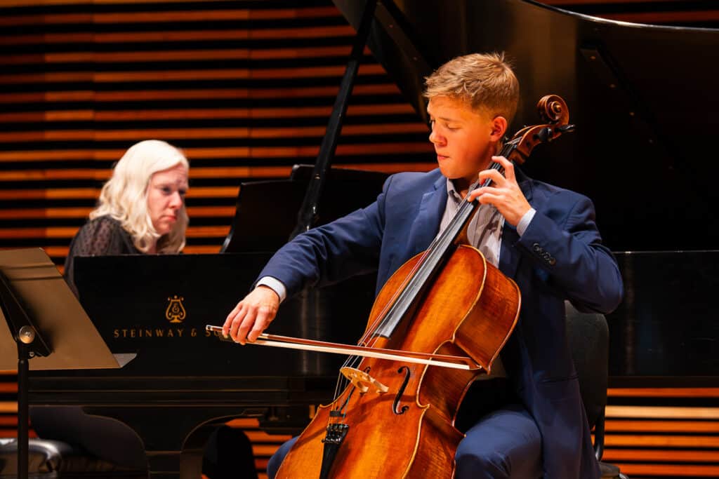 Male student on stage playing cello with female accompanist playing piano in the background.